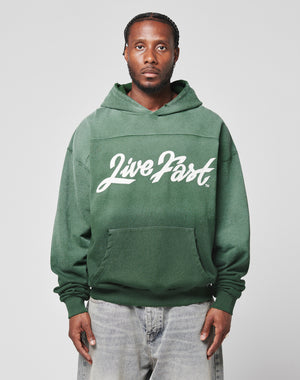 A man with braided hair wears the green LFDY Script Hooded by LIVE FAST DIE YOUNG, featuring Live Fast in white script and Sprayeffekt, paired with light blue Oversize Fit jeans, standing against a gray background.
