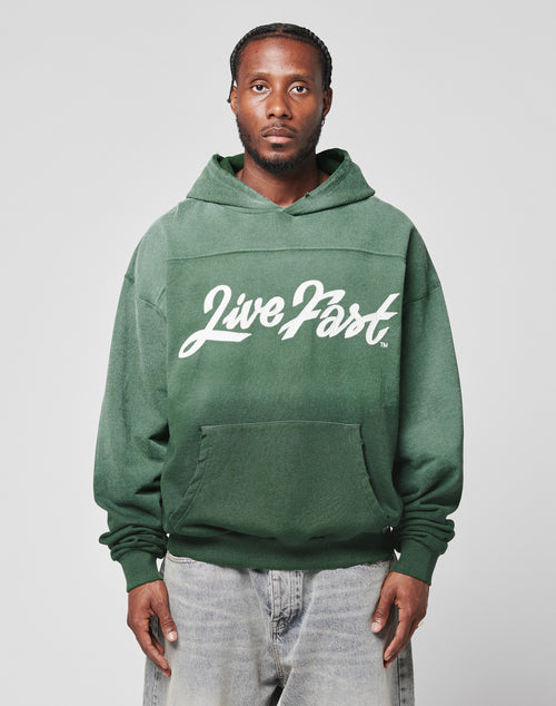 A man with braided hair wears the green LFDY Script Hooded by LIVE FAST DIE YOUNG, featuring Live Fast in white script and Sprayeffekt, paired with light blue Oversize Fit jeans, standing against a gray background.