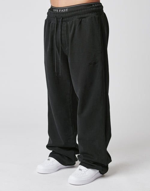 A person in the LIVE FAST DIE YOUNG Basic Wide Jogger—wide fit black sweatpants with an elastic waistband—stands against a plain white background, paired with white sneakers.
