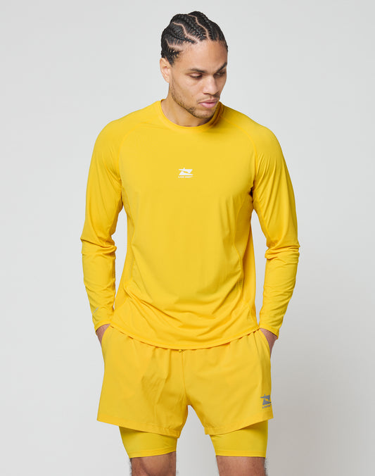 A man wearing the LIVE FAST DIE YOUNG Live Fast Performance Longsleeve and matching yellow shorts with mesh inserts stands with his hands in his pockets, looking down and to the side against a light gray background.
