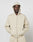 A person in a light beige LIVE FAST DIE YOUNG Basic Script Hooded Zip with the hood up and an oversize fit stands against a white background, hands in the front pockets and wearing a neutral expression.