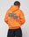 An individual with braided hair faces away, wearing the LIVE FAST DIE YOUNG Basic Orange Hooded made of cotton and polyester, featuring bold front and back “Live Fast Red Orange 17-1464” prints, paired with black pants.