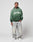 A person stands against a plain background, wearing the LIVE FAST DIE YOUNG LFDY Script Hooded in green with Live Fast Sprayeffekt, light gray distressed baggy jeans, and white sneakers. One hand is in their pocket as they face the camera.