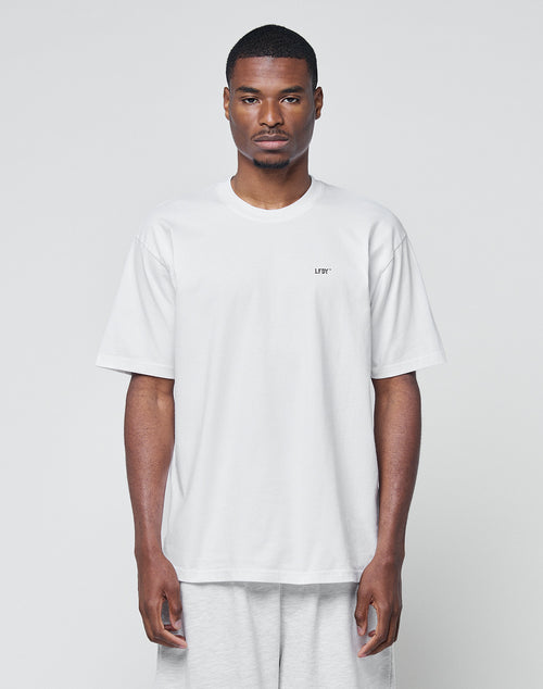 A man stands against a plain light background, wearing LIVE FAST DIE YOUNG’s Basic Chest Tee—a loose-fitting, regular fit white T-shirt with short sleeves and a small LFTP logo print on the left chest—paired with light gray pants.