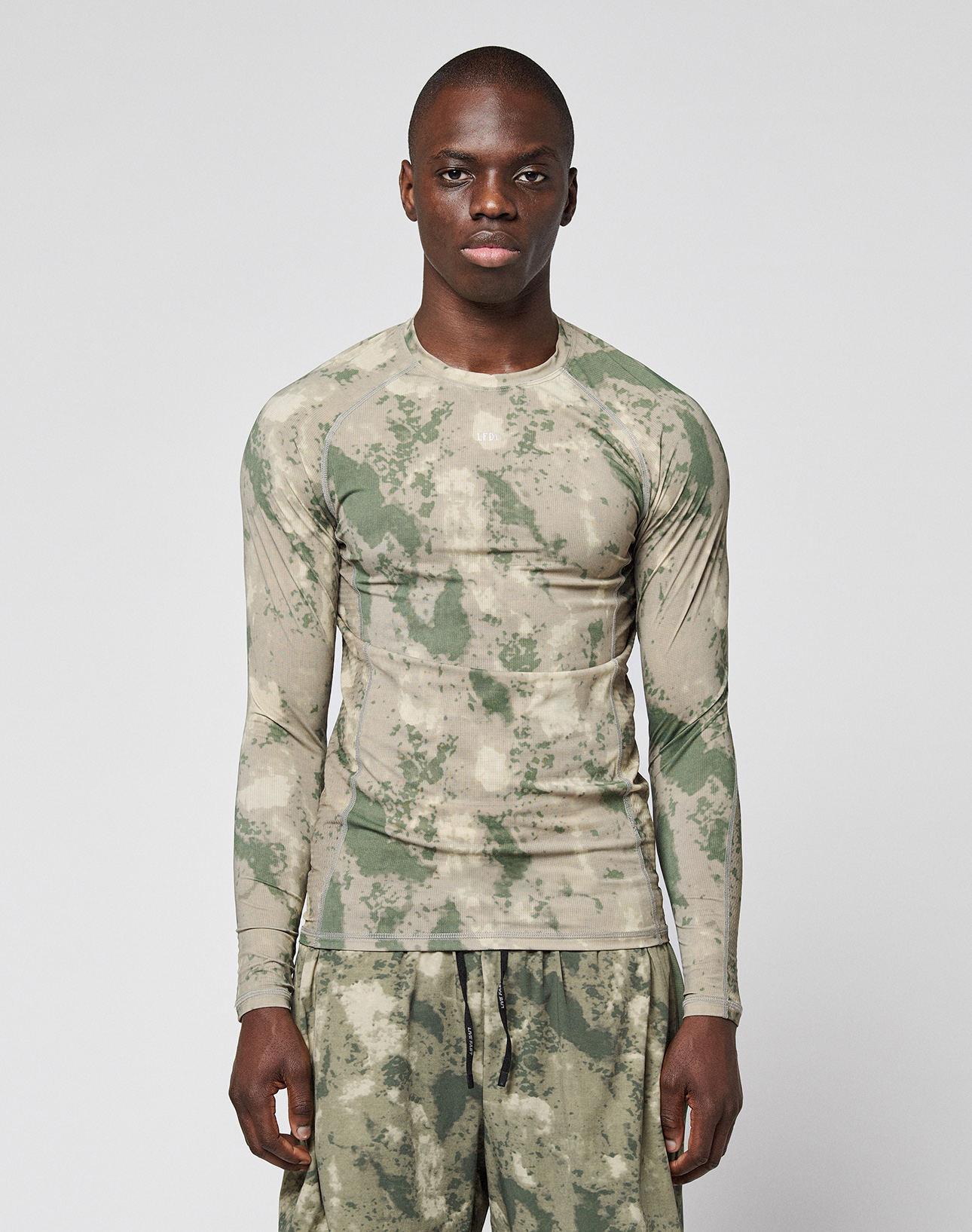 A person wears the LIVE FAST DIE YOUNG LFDY Performance Slim Longsleeve and matching shorts in green and beige camouflage with leichtes Obermaterial, standing against a plain white background.