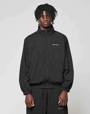 A young man with medium-length, textured hair wears the black LFDY Trackjacket and matching pants from LIVE FAST DIE YOUNG, both featuring subtle Logo-Stick and small LIFES A PARTY text on the left side, standing against a light gray background.