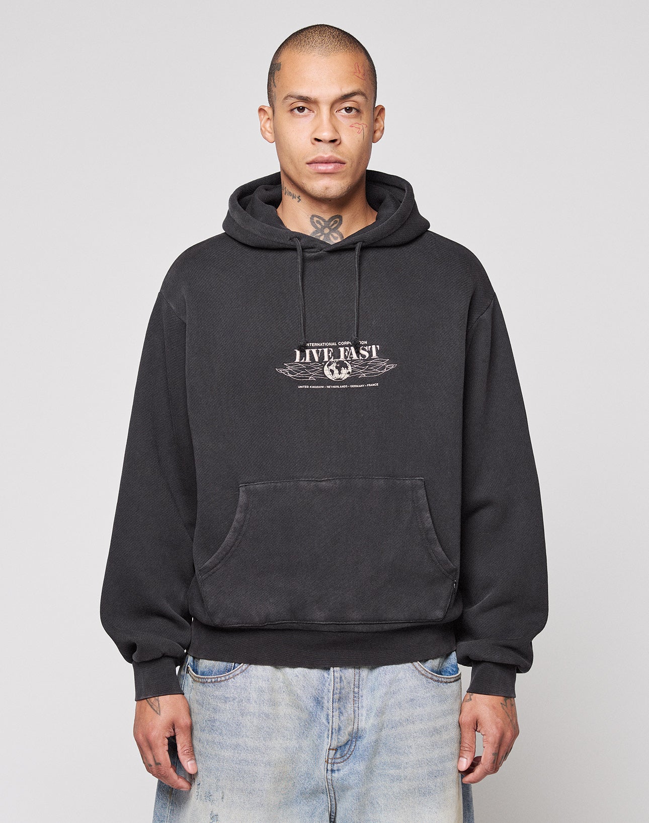 A person wearing a LIVE FAST DIE YOUNG Live Fast Corporation Hooded sweatshirt in black with faded artwork stands against a light gray background, paired with light blue jeans, short hair, and visible neck tattoos.