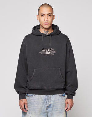 A person wearing a LIVE FAST DIE YOUNG Live Fast Corporation Hooded sweatshirt in black with faded artwork stands against a light gray background, paired with light blue jeans, short hair, and visible neck tattoos.