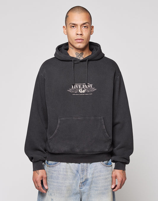 A person wearing a LIVE FAST DIE YOUNG Live Fast Corporation Hooded sweatshirt in black with faded artwork stands against a light gray background, paired with light blue jeans, short hair, and visible neck tattoos.