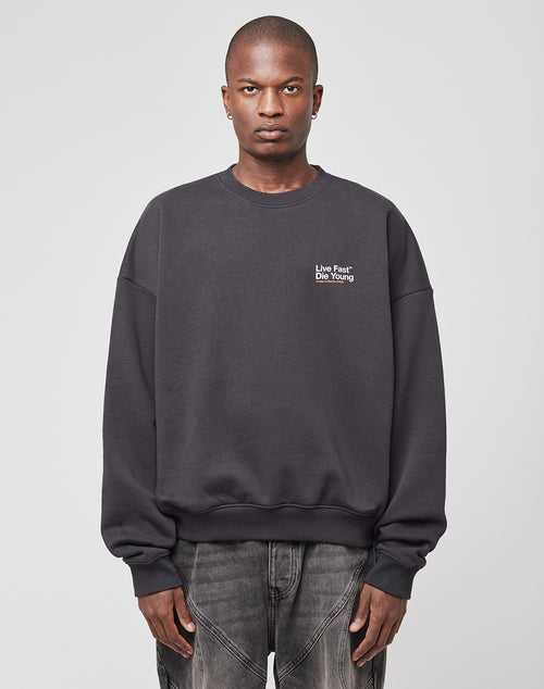 A person faces the camera in a dark gray oversized Live Fast NY Sweater by LIVE FAST DIE YOUNG, featuring Live Fast Die Young text on the upper left chest, paired with faded jeans against a plain light background.