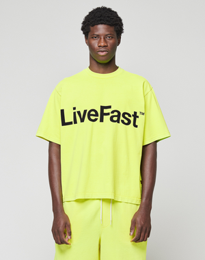 A person wears the LIVE FAST DIE YOUNG Basic OS Livefast Tee in neon yellow, featuring an oversize fit and bold "LiveFast™" black front print, paired with matching shorts against a plain background. Made from 100% Baumwolle.