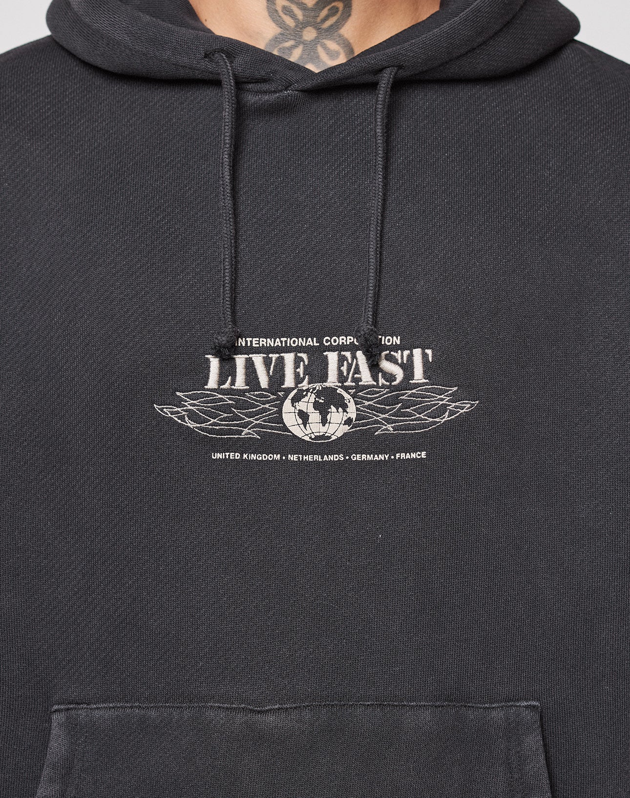 Close-up of a person wearing the Live Fast Corporation Hooded by LIVE FAST DIE YOUNG, a black oversized hoodie featuring a LIVE FAST print and globe design on the chest, plus country names: United Kingdom, Netherlands, Germany, France. Neck tattoo visible.