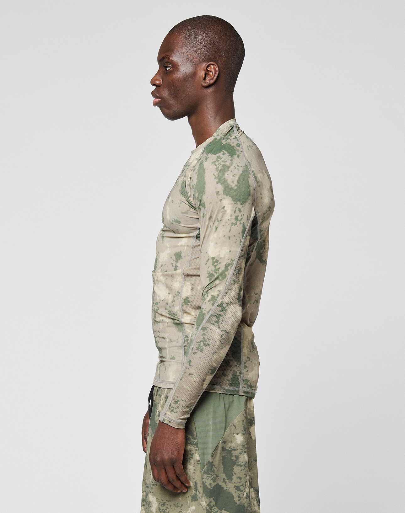A man stands in profile against a white background, wearing the LIVE FAST DIE YOUNG LFDY Performance Slim Longsleeve and matching Slim Fit pants in green and beige camouflage, both crafted from leichtes Obermaterial.