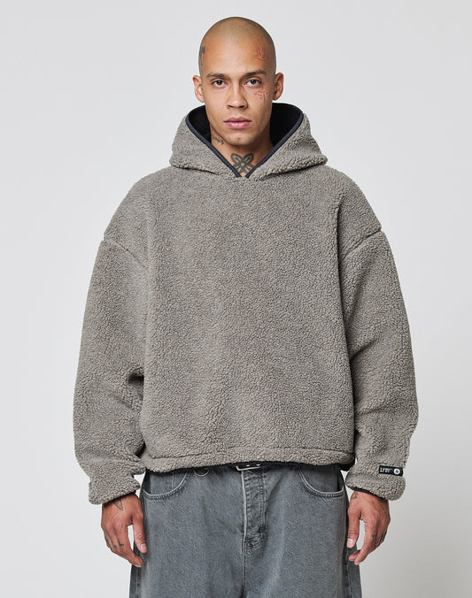A person with a shaved head wears the LIVE FAST DIE YOUNG LFDY Teddy Hooded in gray, featuring practical Eingrifftaschen, and loose-fitting gray jeans, standing against a plain white background.