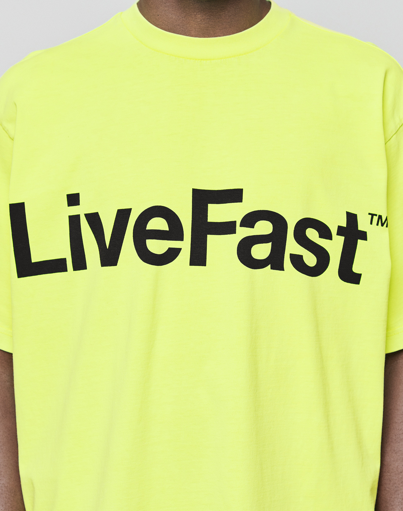 A person is seen in the Basic OS Livefast Tee by LIVE FAST DIE YOUNG, featuring an oversized fit and bold "LiveFast™" black front print on bright yellow 100% cotton. The person's face is not shown.