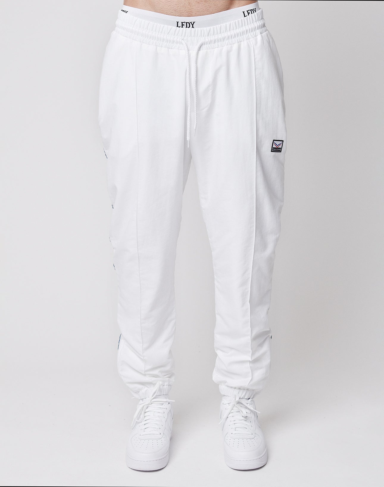 A person wears LIVE FAST DIE YOUNG LFDY x Trigema Trackpants in regular fit with a drawstring waist and a small rectangular patch on the left thigh, paired with white sneakers against a plain light background.