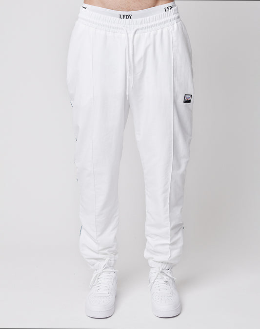A person wears LIVE FAST DIE YOUNG LFDY x Trigema Trackpants in regular fit with a drawstring waist and a small rectangular patch on the left thigh, paired with white sneakers against a plain light background.