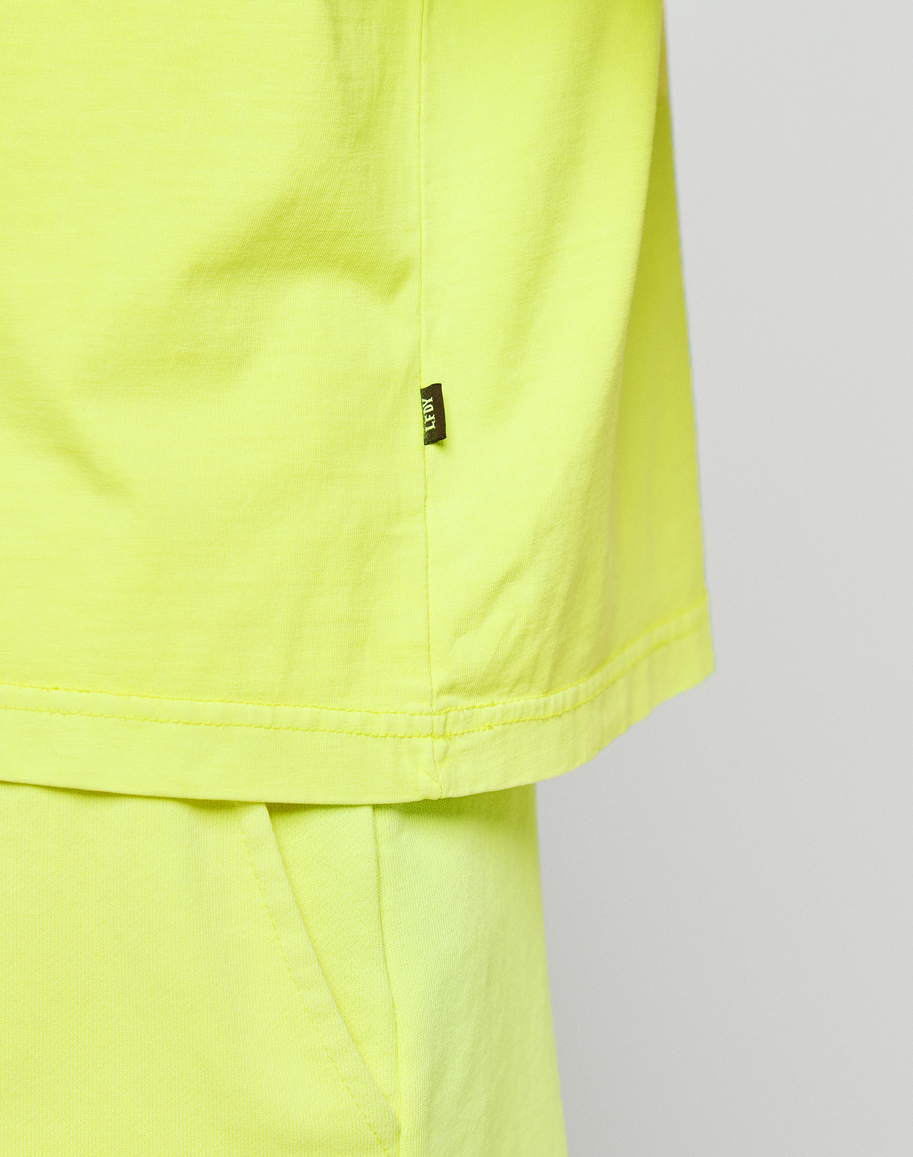 Close-up of a person wearing LIVE FAST DIE YOUNG’s bright yellow Basic OS Livefast Tee, showing the side with a small black tag and the top of matching pants featuring a pocket.