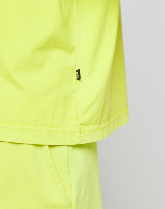 Close-up of a person wearing LIVE FAST DIE YOUNG’s bright yellow Basic OS Livefast Tee, showing the side with a small black tag and the top of matching pants featuring a pocket.