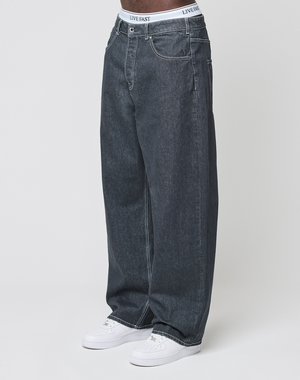 A person wears LFDY Baggy Denim by LIVE FAST DIE YOUNG, pairing the dark wash jeans with white sneakers and a visible waistband reading LIVE FAST, against a plain white background.