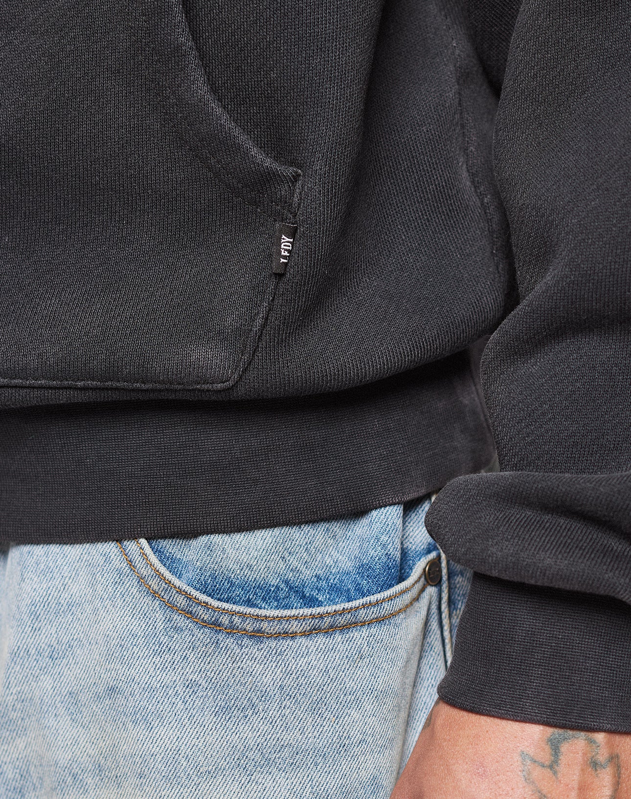 Close-up of a person wearing the LIVE FAST DIE YOUNG Live Fast Corporation Hooded in black, oversized fit, with a small fabric tag on the pocket, paired with light blue denim jeans. A tattoo on their wrist is partially visible.