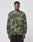 A man stands facing forward in LIVE FAST DIE YOUNGs Copyright Waffle Longsleeve, an oversize fit camo shirt with bold back print, paired with black pants, set against a plain light gray background.