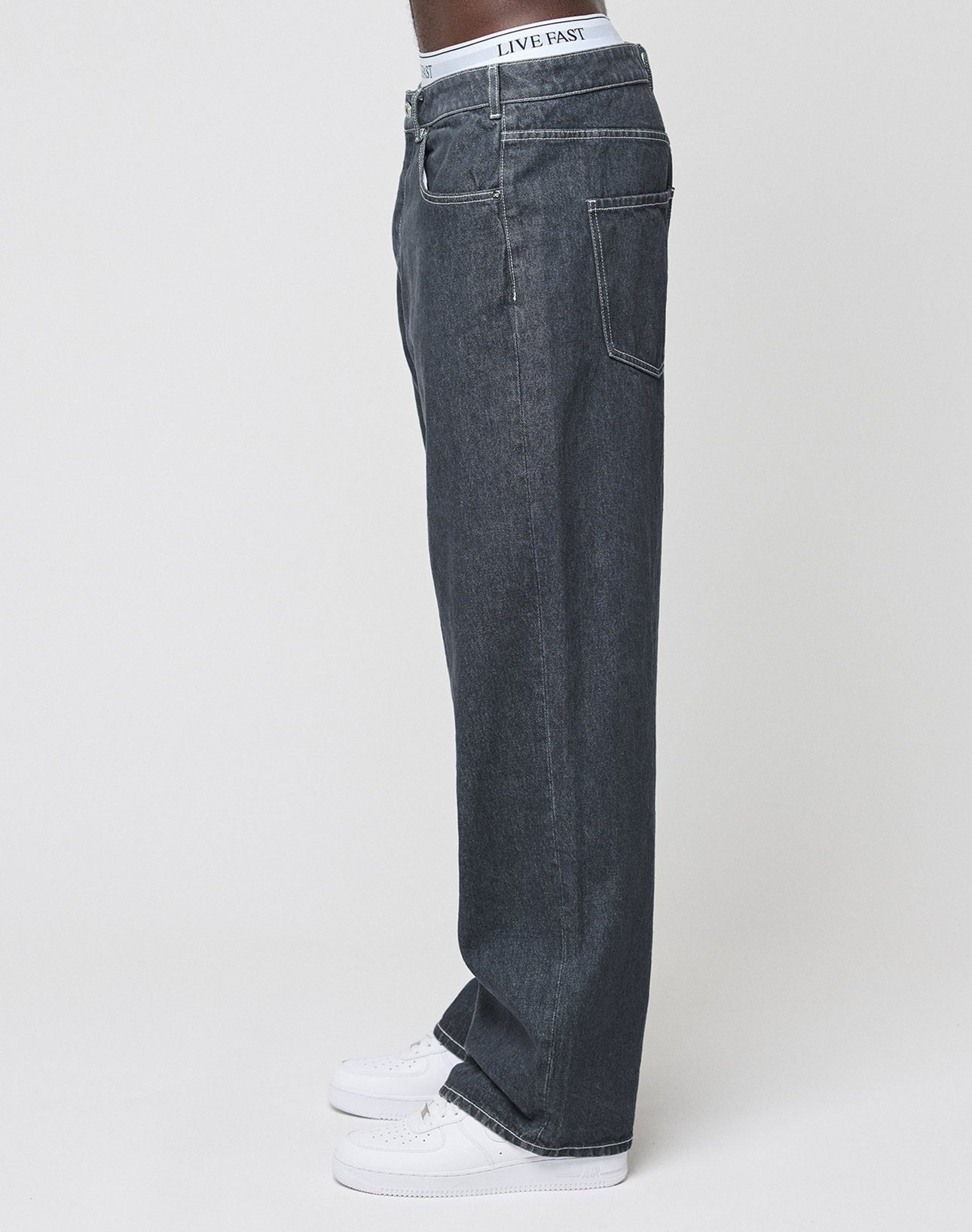 A person in profile wears LIVE FAST DIE YOUNG’s LFDY Baggy Denim jeans (100% Baumwolle), white sneakers, and a waistband with "LIVE FAST" text. The background is plain and light-colored.