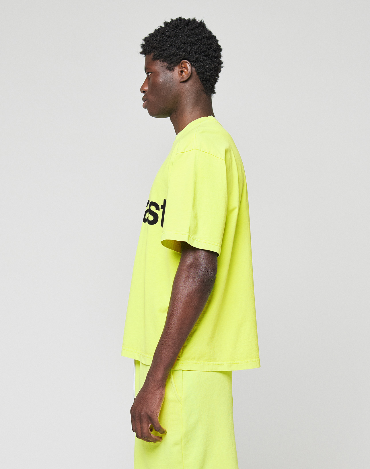 A person stands in profile against a plain background, wearing the LIVE FAST DIE YOUNG Basic OS Livefast Tee—an oversize, short-sleeved neon yellow 100% Baumwolle shirt with a partially visible black frontprint—and matching shorts.