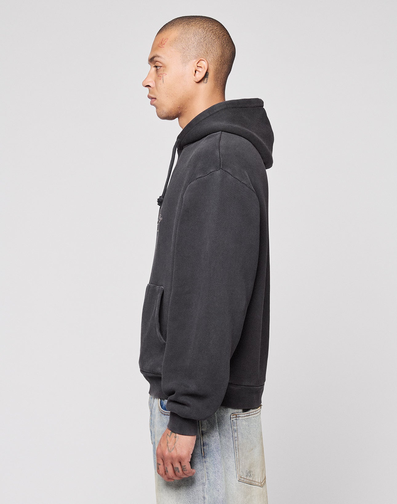 A person with short hair stands in profile against a plain background, wearing the LIVE FAST DIE YOUNG Live Fast Corporation Hooded sweatshirt and light-wash jeans in Vintage Waschung, hands relaxed by their side.