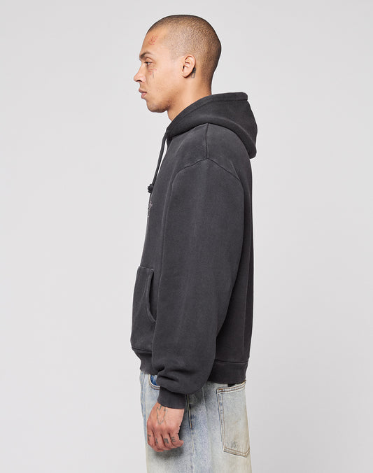 A person with short hair stands in profile against a plain background, wearing the LIVE FAST DIE YOUNG Live Fast Corporation Hooded sweatshirt and light-wash jeans in Vintage Waschung, hands relaxed by their side.