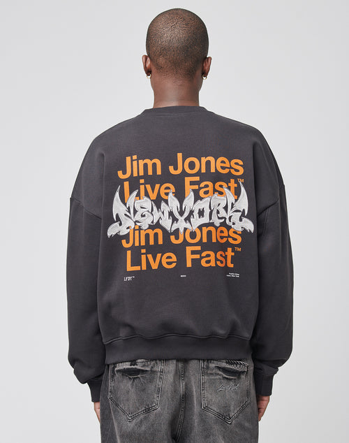 A person in the LIVE FAST DIE YOUNG Live Fast NY Sweater—an oversized black sweatshirt with orange Jim Jones Live Fast front and back print plus white graffiti-style New York text—paired with faded black jeans. Cotton-polyester blend. Viewed from the back.
