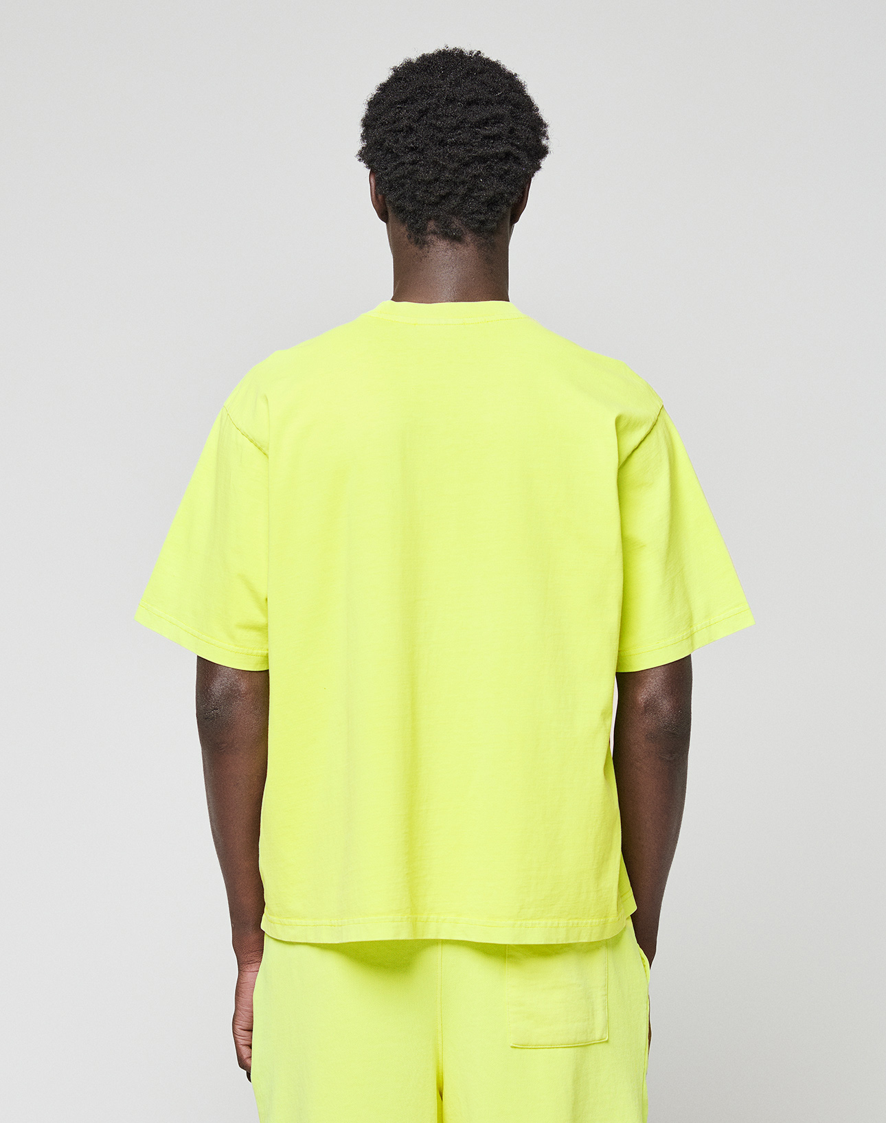 A person with short curly hair faces away from the camera, wearing a bright yellow LIVE FAST DIE YOUNG Basic OS Livefast Tee and matching shorts against a plain, light background.