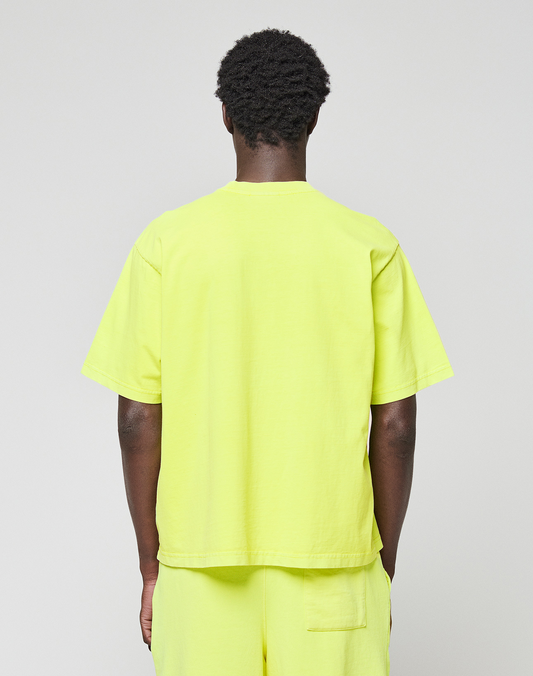 A person with short curly hair faces away from the camera, wearing a bright yellow LIVE FAST DIE YOUNG Basic OS Livefast Tee and matching shorts against a plain, light background.