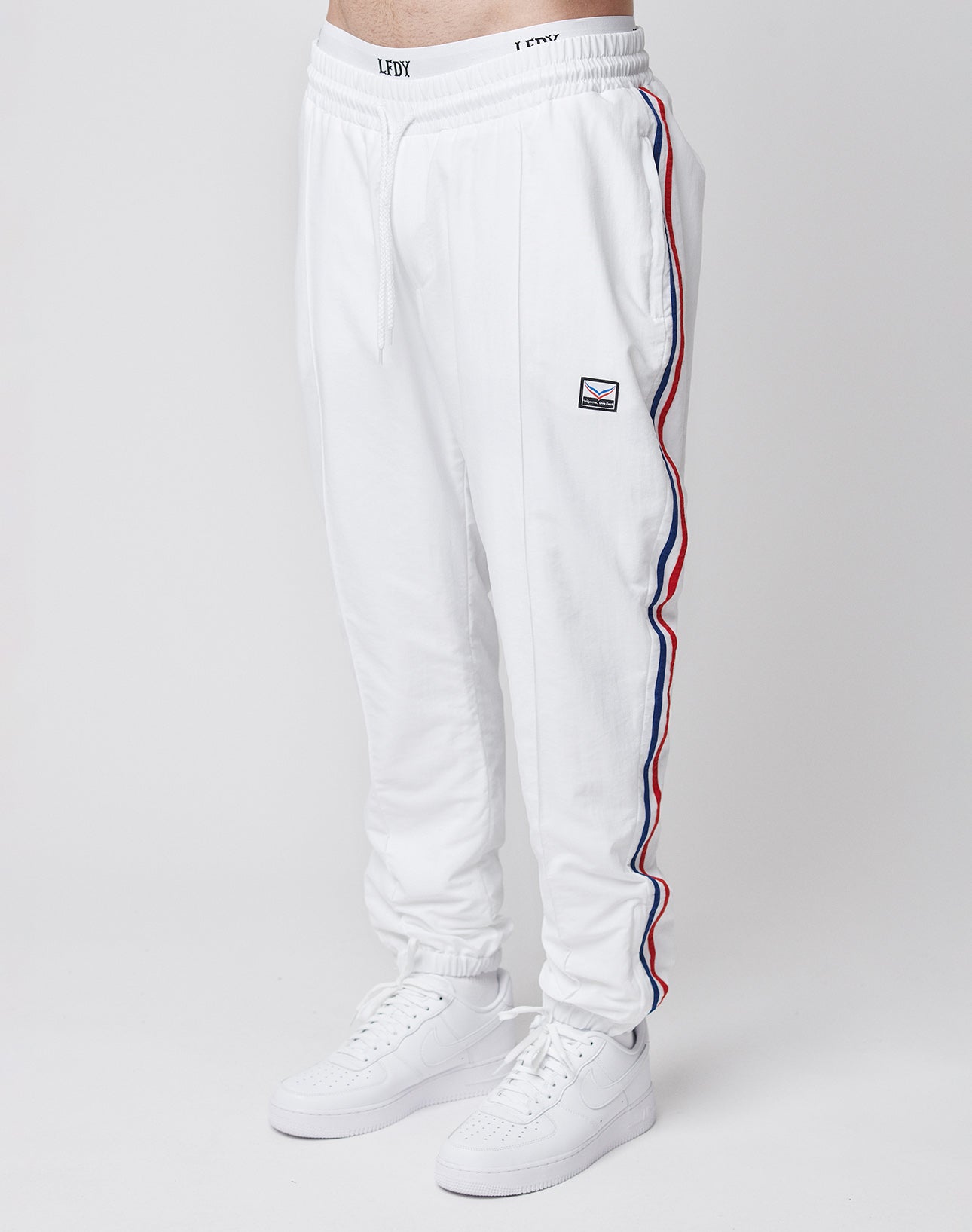 A person wears LIVE FAST DIE YOUNG LFDY x Trigema Trackpants—regular fit, white with red and blue side stripes, a small patch on the left thigh—and white sneakers, standing against a plain white background.