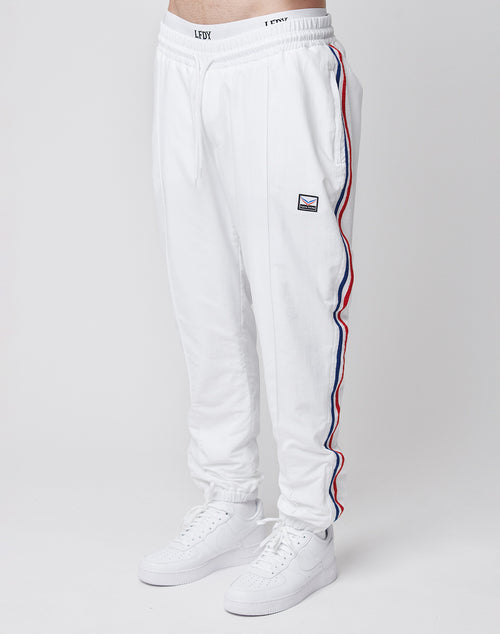 A person wears LIVE FAST DIE YOUNG LFDY x Trigema Trackpants—regular fit, white with red and blue side stripes, a small patch on the left thigh—and white sneakers, standing against a plain white background.