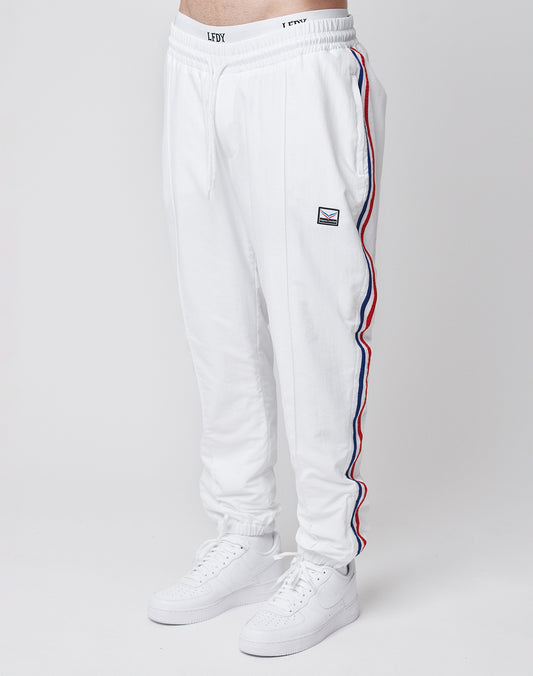 A person wears LIVE FAST DIE YOUNG LFDY x Trigema Trackpants—regular fit, white with red and blue side stripes, a small patch on the left thigh—and white sneakers, standing against a plain white background.