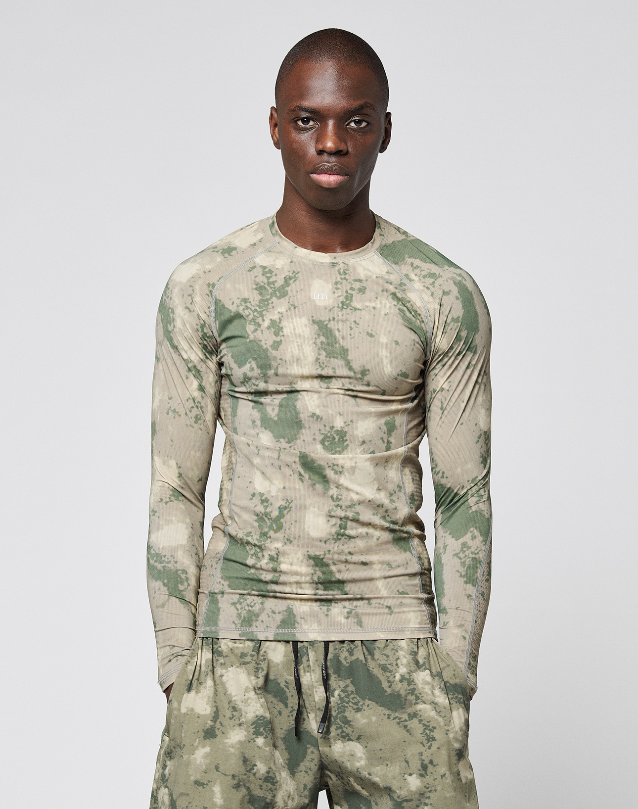 A person faces forward, wearing the LIVE FAST DIE YOUNG LFDY Performance Slim Longsleeve with camouflage pattern, reflektierender Logo Print, and matching lightweight shorts against a plain light background.