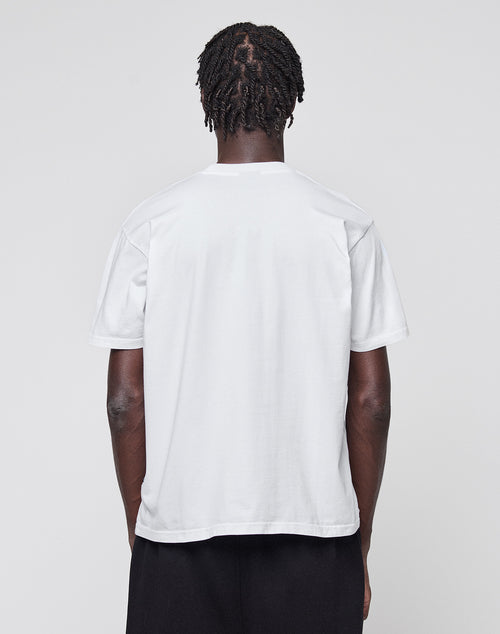 A person with short, twisted hair stands with their back to the camera, wearing a plain white LIVE FAST DIE YOUNG Abus Magomedov Tee and black pants against a light gray background.