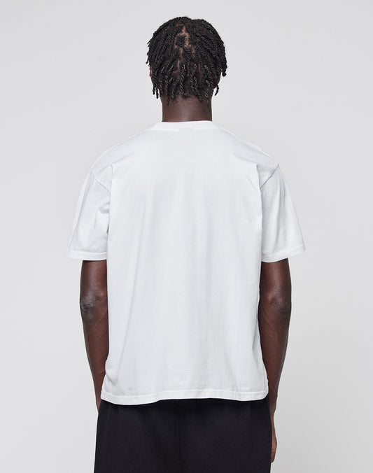 A person with short, twisted hair stands with their back to the camera, wearing a plain white LIVE FAST DIE YOUNG Abus Magomedov Tee and black pants against a light gray background.