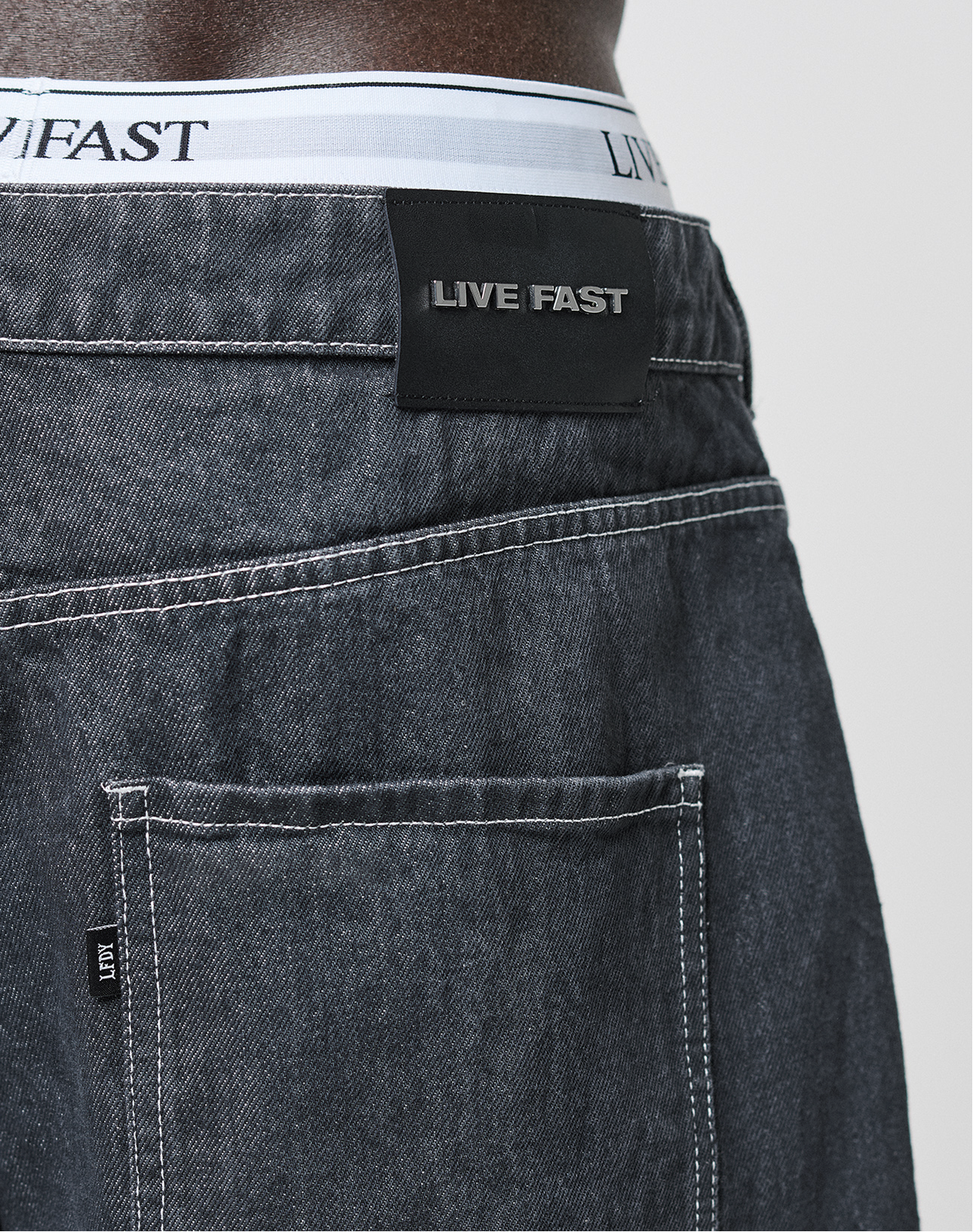 Close-up of the back of LFDY Baggy Denim jeans by LIVE FAST DIE YOUNG in dark gray with white stitching, a black LIVE FAST label above the pocket, and white underwear waistband showing LIVE FAST text. Made from 100% Baumwolle.