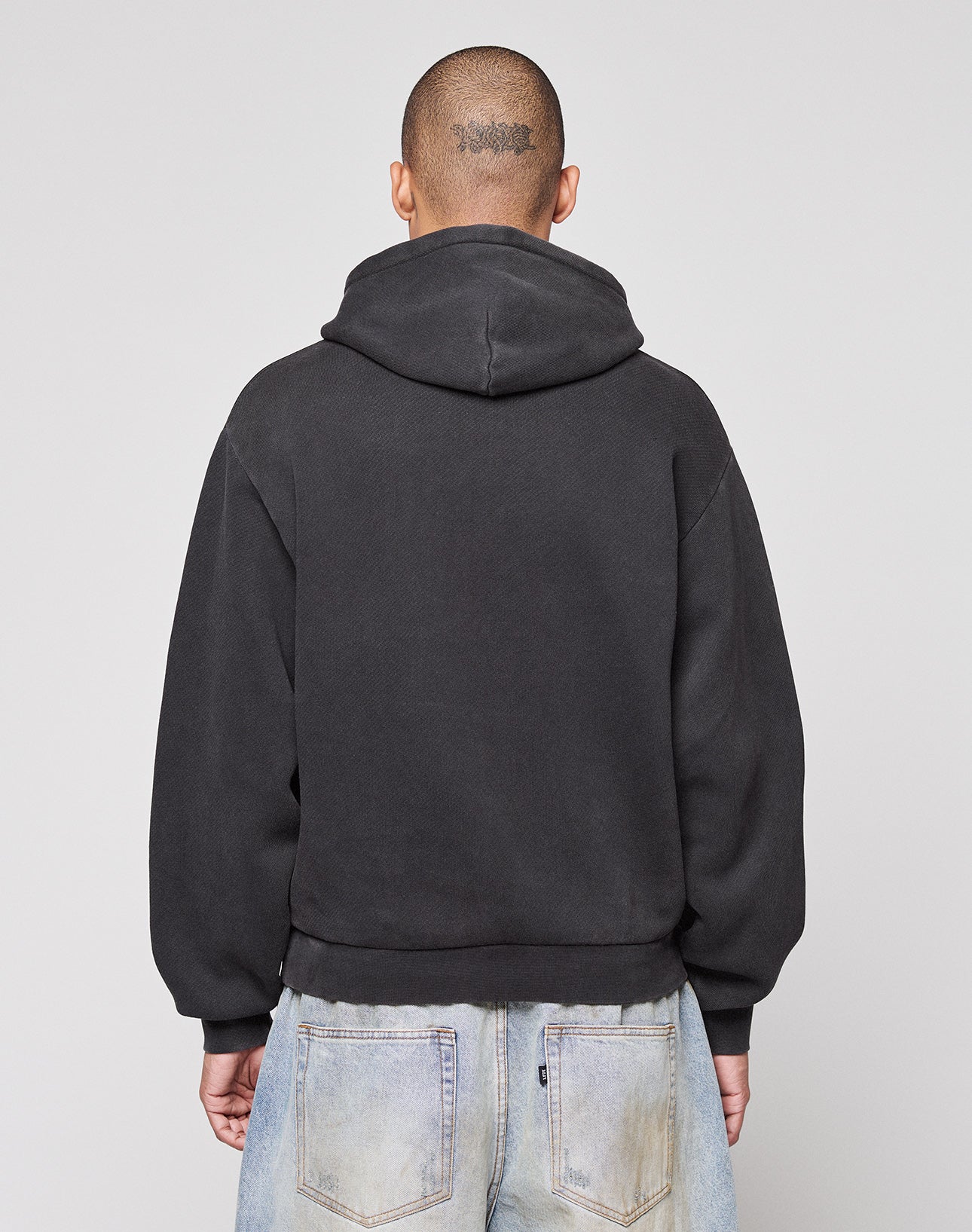 A person with a shaved head and a tattoo is wearing the LIVE FAST DIE YOUNG Live Fast Corporation Hooded in black with print artwork and vintage-washed light blue jeans, standing with their back to the camera against a plain light background.