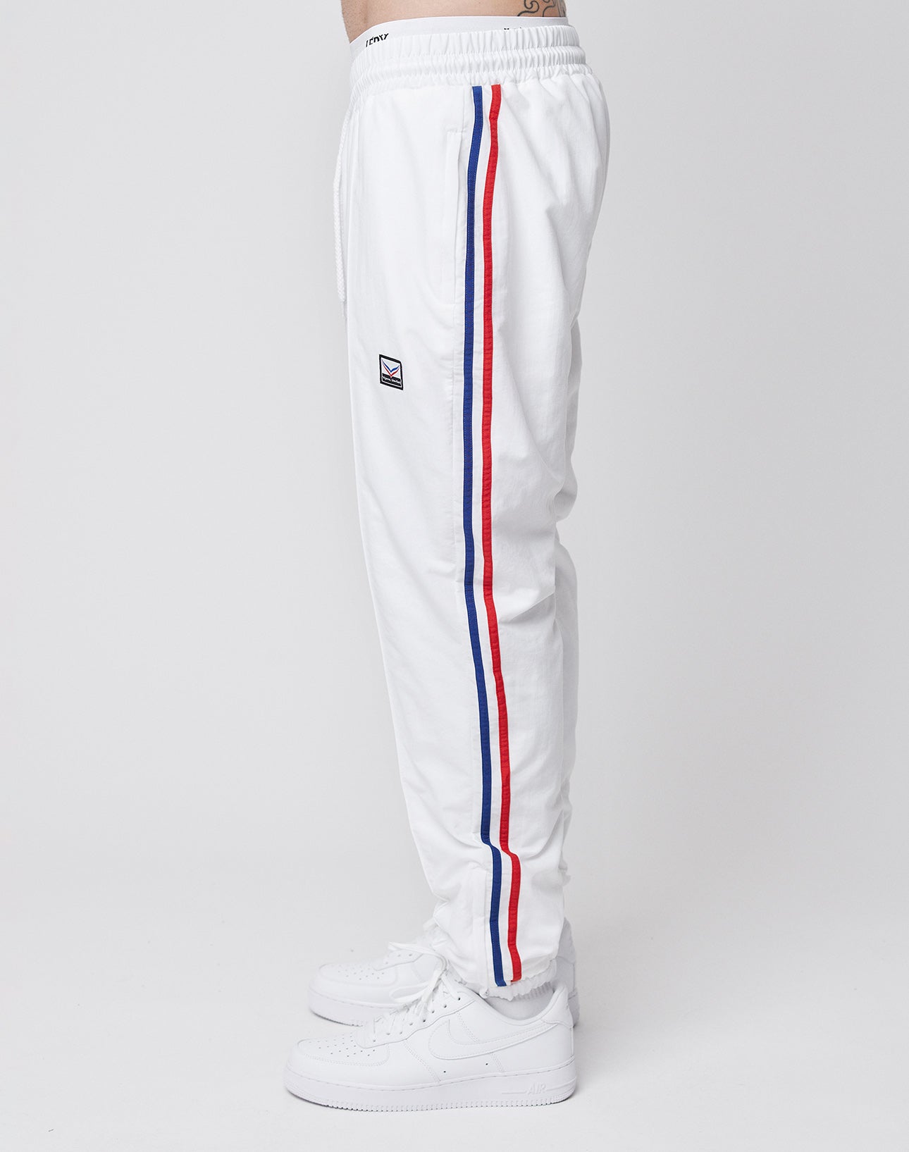 A person in LIVE FAST DIE YOUNG LFDY x Trigema Trackpants—white with red and blue side stripes, a small embroidered thigh logo, and white sneakers—stands sideways against a plain white background.