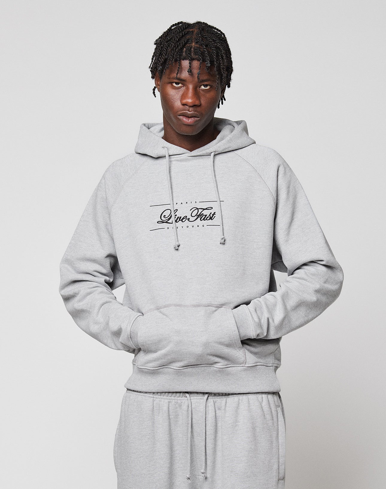 A person with short braided hair and a neutral expression models the light gray LFDY Embroidery Hooded by LIVE FAST DIE YOUNG, featuring “Live Fast” script on the front and matching sweatpants, against a plain white background.