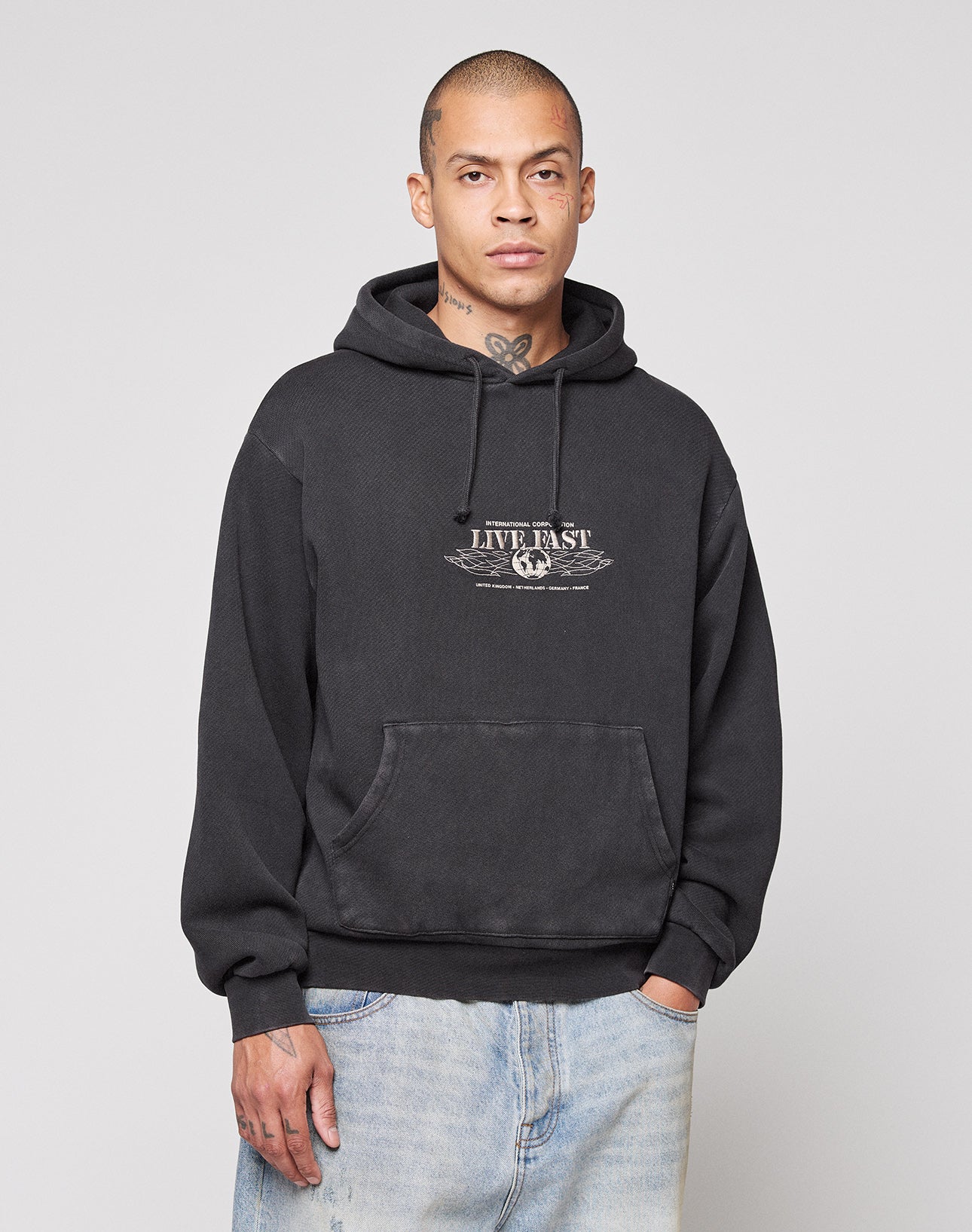 A person with a shaved head and visible tattoos wears the LIVE FAST DIE YOUNG Live Fast Corporation Hooded, an oversized black sweatshirt with “LIVE FAST” artwork, paired with light blue jeans against a plain light gray background.