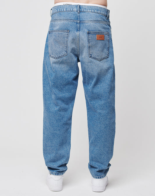 A person, seen from the back against a plain white background, wears LIVE FAST DIE YOUNG Regular Denim jeans with two back pockets, subtle branding, a brown leather patch above the right pocket, and white sneakers.