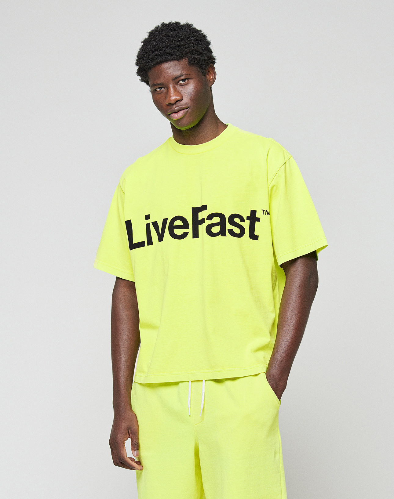 A man stands against a plain background wearing the LIVE FAST DIE YOUNG Basic OS Livefast Tee, an oversized bright yellow T-shirt made from 100% Baumwolle with "LiveFast" printed in large black letters on the front.