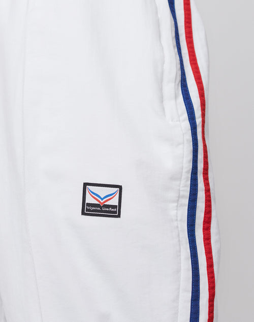 Close-up of LFDY x Trigema Trackpants by LIVE FAST DIE YOUNG: white athletic pants with red and blue side stripes, a small rectangular patch near the pocket with stylized Epimena. Live Fast text. Limitierte Auflage, Regular Fit.