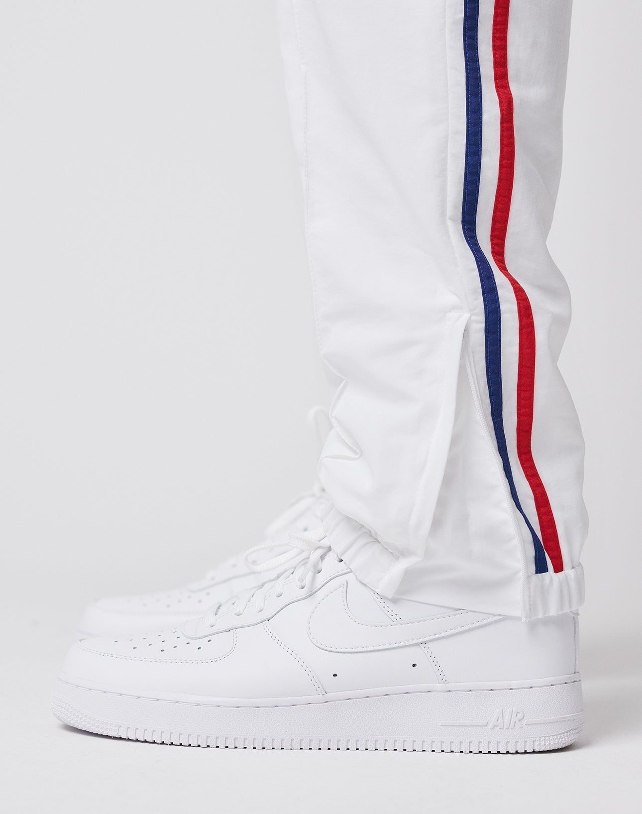 Someone wears LIVE FAST DIE YOUNG LFDY x Trigema Trackpants—white with red and blue side stripes, regular fit—paired with white Nike Air Force 1 sneakers, standing against a plain white background.