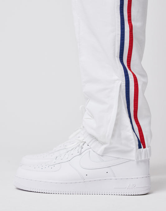 Someone wears LIVE FAST DIE YOUNG LFDY x Trigema Trackpants—white with red and blue side stripes, regular fit—paired with white Nike Air Force 1 sneakers, standing against a plain white background.
