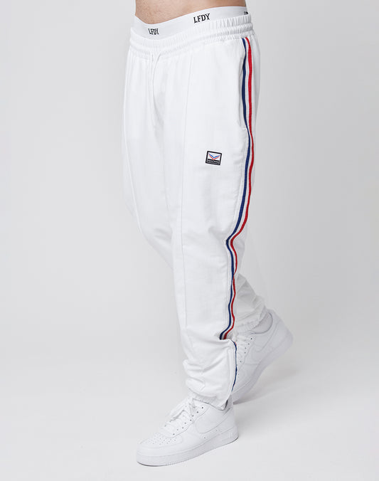 A person wears LIVE FAST DIE YOUNG LFDY x Trigema Trackpants in white with red and blue side stripes, a small rectangular logo on the upper leg, regular fit, paired with white sneakers, standing against a plain white background.